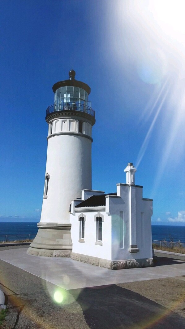 Un faro bianco con una lanterna in vetro, una struttura secondaria adiacente e un cielo azzurro con luce solare.
