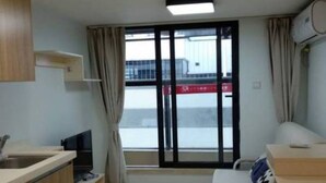 Ein kleines Hotelzimmer mit Küchenzeile, einem Esstisch, einem Fernseher und einem großen Fenster mit Vorhängen.