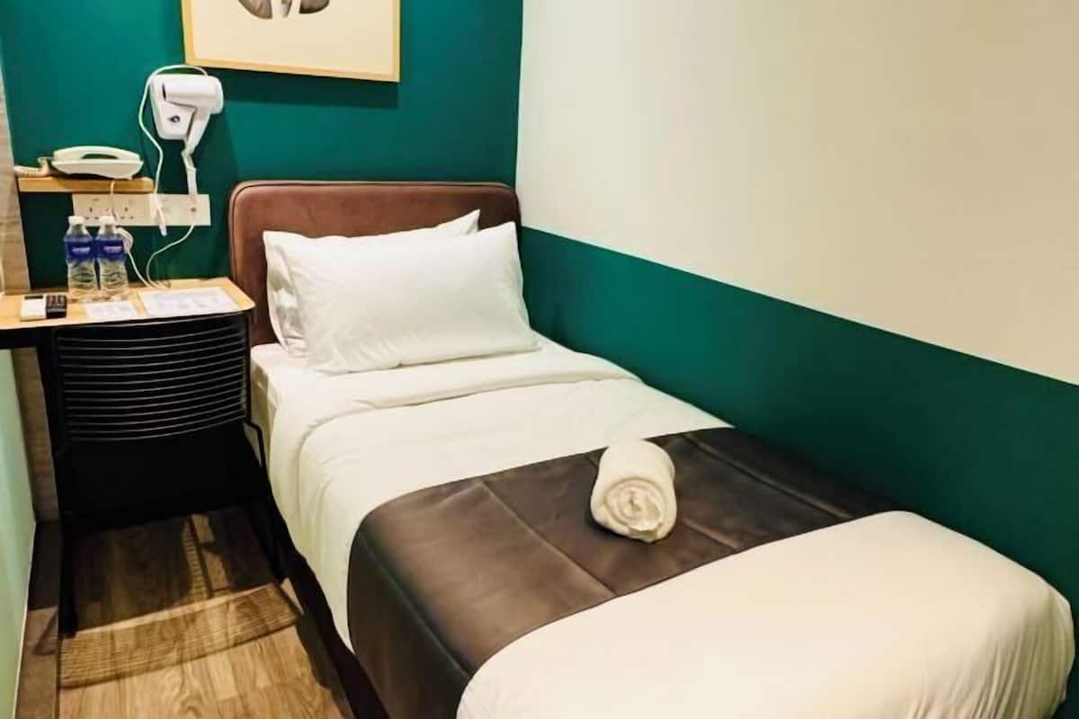 A hotel room with a bed, a bedside table with a phone and water bottles, a wall-mounted hairdryer, and a framed picture above the bed.