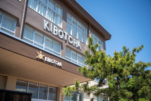 A hotel named "KIBOTCHA" with a modern facade, large windows, and a sign with a sun icon.