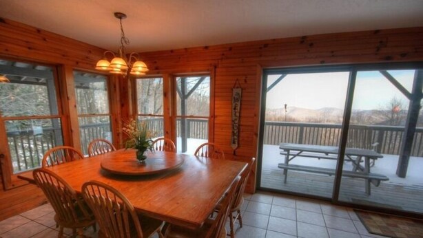 A wooden dining room with a large table and chairs, a hanging light fixture, and a view of a deck and mountains through large windows. - Mountain Top Private Retreat (todd)