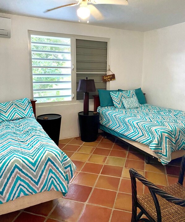 Une chambre avec deux lits, un ventilateur de plafond, une fenêtre avec des stores, une lampe et une poubelle.