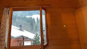 L’intérieur d’un chalet en bois, avec une grande fenêtre, un luminaire suspendu et une vue sur un paysage enneigé.
