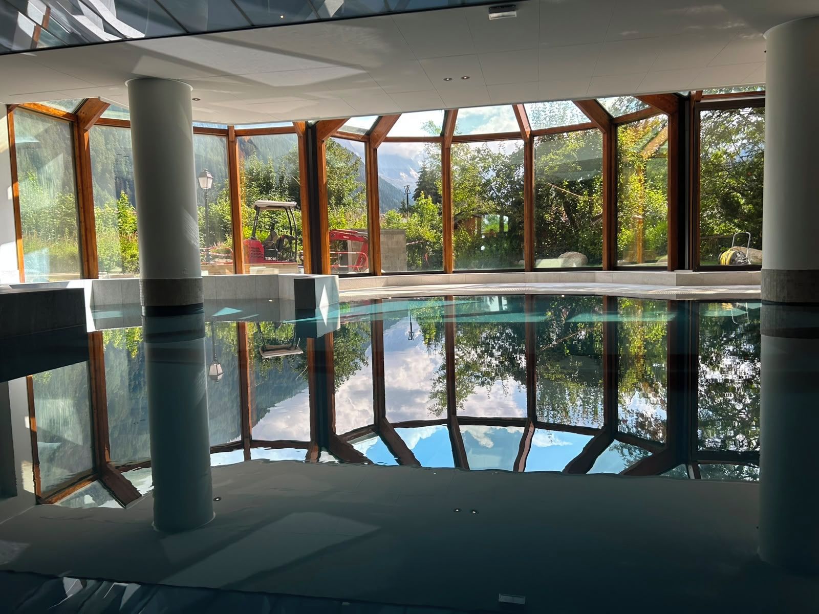 An indoor swimming pool with clear glass walls, wooden beams, and a view of greenery outside.