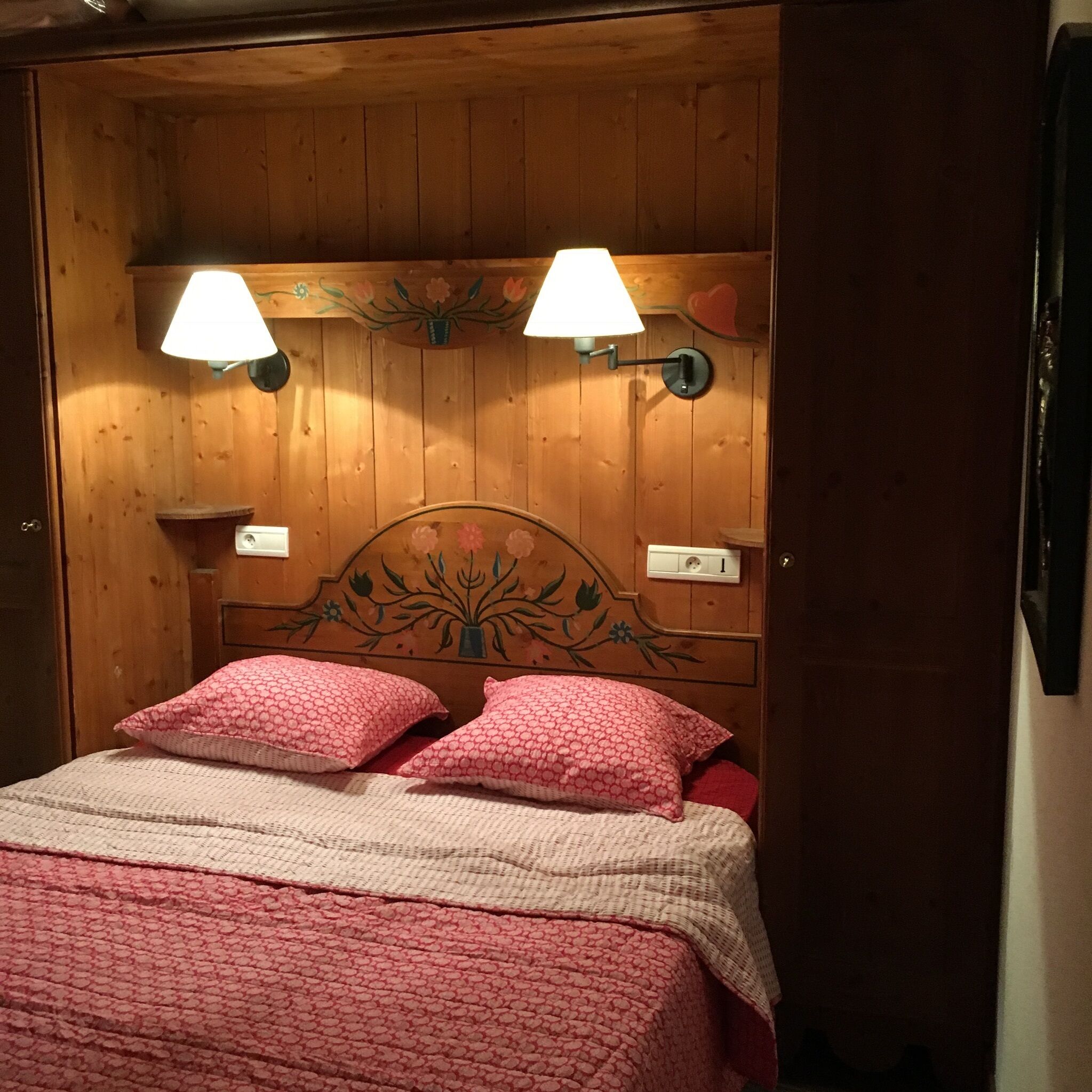 A wooden room with a bed, two wall-mounted lamps, and a floral headboard.