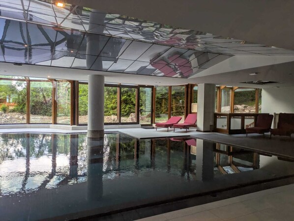 Une piscine intérieure aux parois en verre transparent, dotée de chaises longues rouges et offrant une vue sur la verdure environnante.