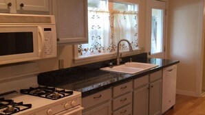 Una cocina con armarios blancos, microondas, estufa, lavaplatos y una ventana con cortinas de flores.