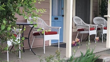 Relax on the porch with comfortable wicker chairs and a small table, surrounded by lush greenery and a charming entrance.