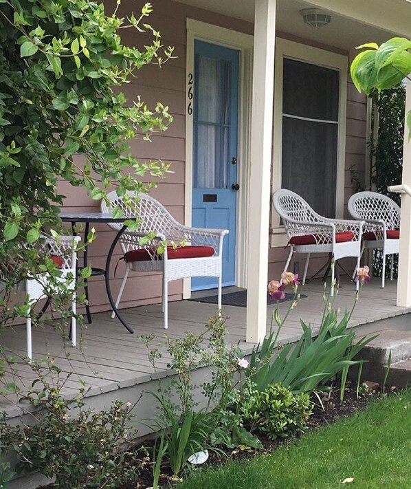 Relax on the porch with comfortable wicker chairs and a small table, surrounded by lush greenery and a charming entrance.