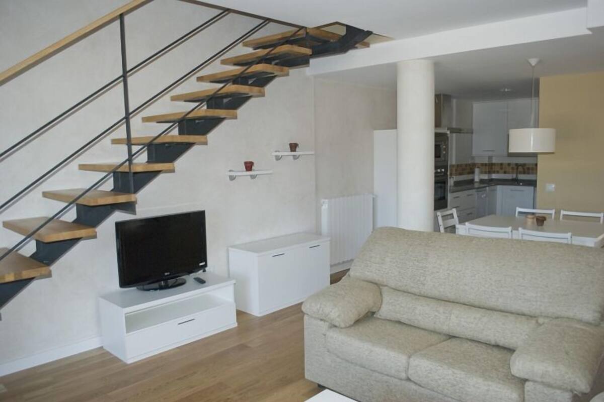 Un salon moderne avec un escalier en bois, un téléviseur à écran plat posé sur un meuble blanc, un canapé beige et, au fond, un espace cuisine.