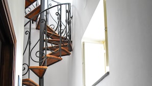 Un escalier en colimaçon avec des marches en bois et des rampes en métal noir, situé dans un couloir aux murs blancs et au sol carrelé.
