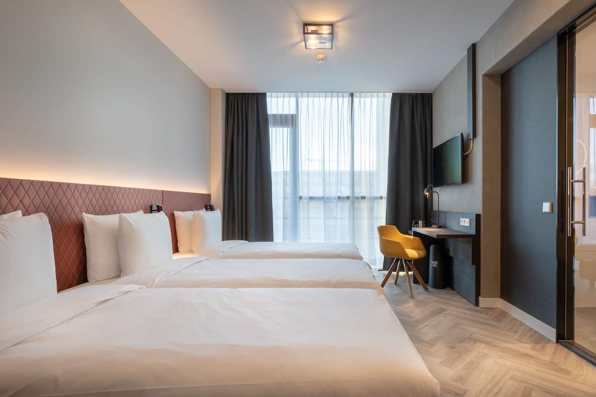 A hotel room with two beds, a desk, a chair, a TV, and a large window with curtains.