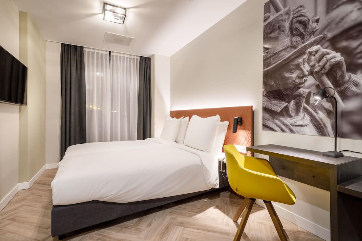 A modern hotel room with a large bed, a desk with a chair, a flat-screen TV, and a large artwork on the wall.