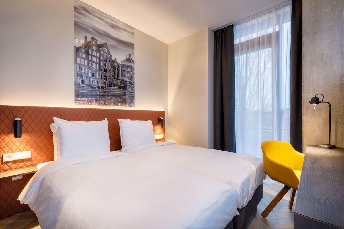 A hotel room with a large bed, a yellow chair, a painting of buildings, and a window with curtains.