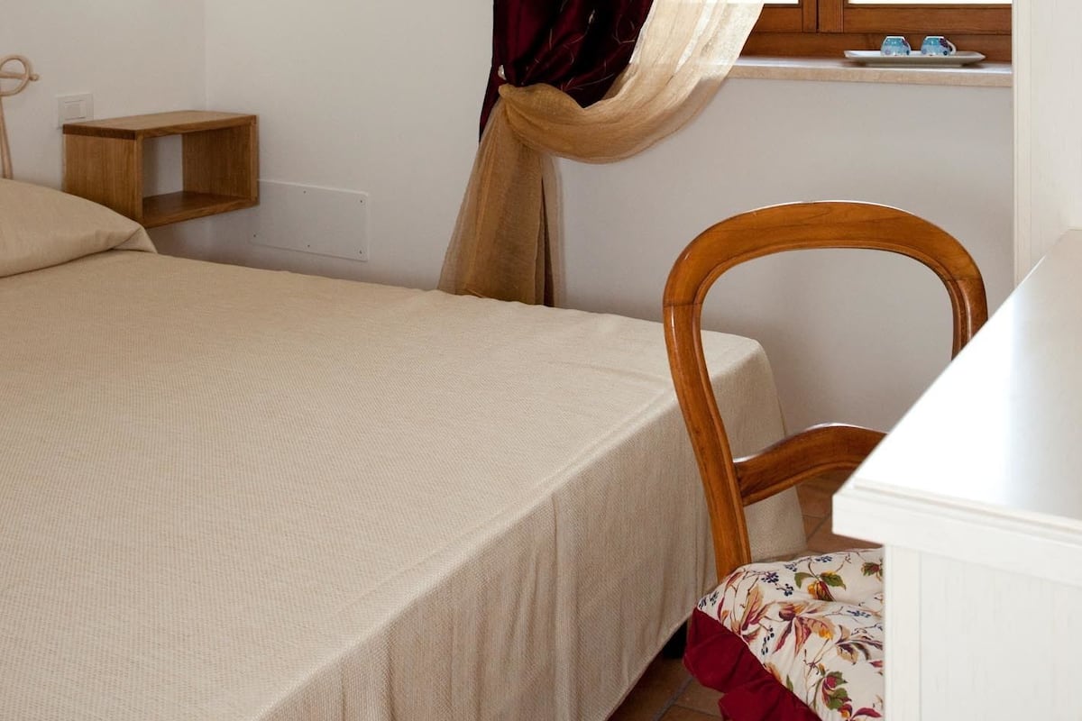 A bedroom with a bed, a wooden chair, a nightstand, a wall-mounted lamp, and a window with curtains.