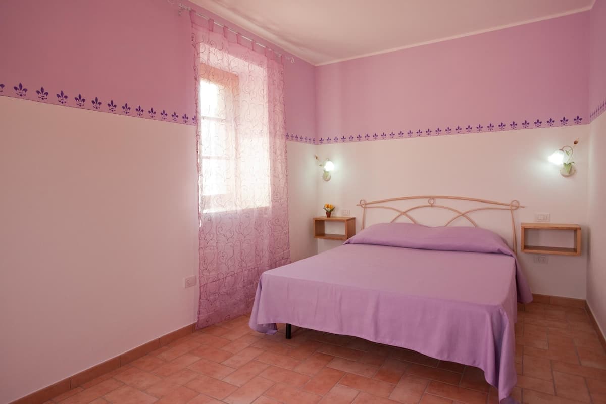 A bedroom with a bed, a nightstand, a wall-mounted lamp, and a window with a curtain.