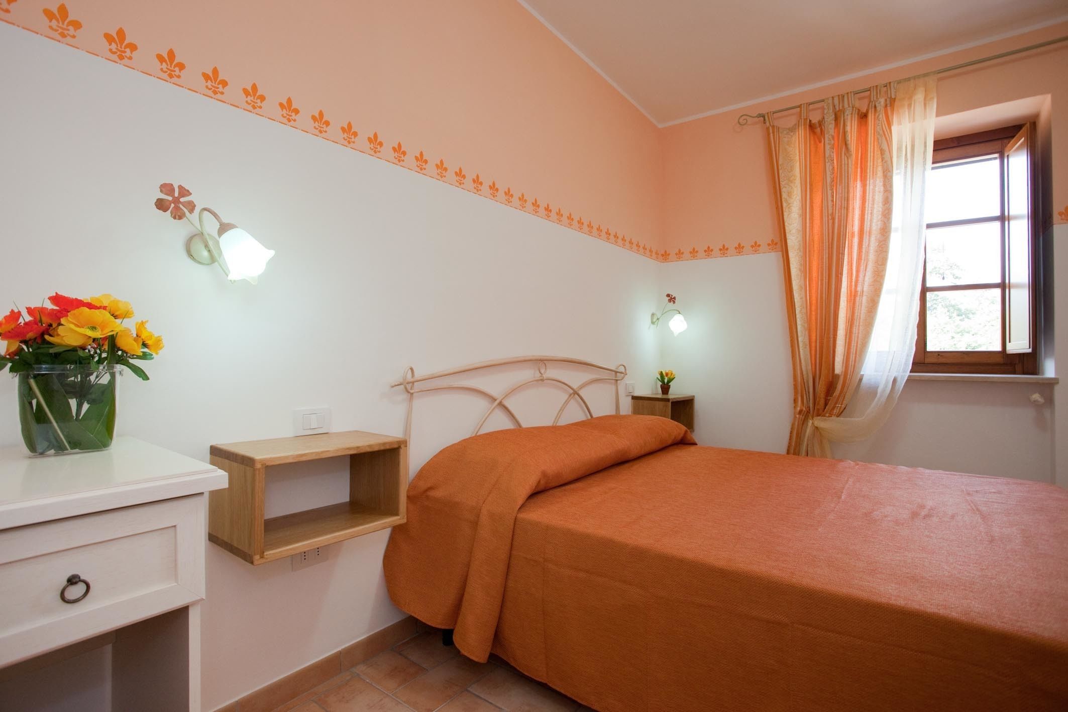 A bedroom with a bed, a nightstand, a lamp, a vase with flowers, and a window with curtains.