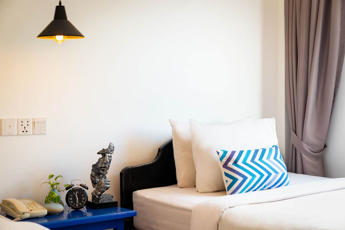 Une chambre d’hôtel bien rangée, avec un lit, une table de chevet dotée d’une lampe, un téléphone, une plante et une horloge murale.