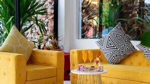 A yellow sofa with patterned cushions, a small round table with drinks and a decorative tray, and a view of a stone wall and greenery through large windows. - Popular Hotel & Spa (Siem Reap)