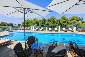 Piscina con sillones de descanso y sombrillas, rodeada de una cerca y árboles.
