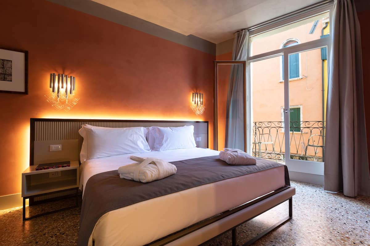 A hotel room with a large bed, a nightstand, a wall-mounted lamp, and a view of a building through a window.