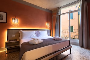 Habitación de hotel con una cama grande, una mesita de noche, una lámpara de pared y vista a un edificio a través de una ventana.