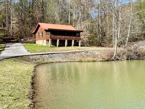 Un chalet rustique avec un toit rouge, situé au bord d’un étang, entouré d’arbres.