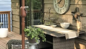 A wooden deck with a round table set for dining, a clock on the wall, and a potted plant.