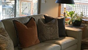 A living room with a beige sofa, patterned cushions, a black throw blanket, a side table with a lamp, and a potted plant.