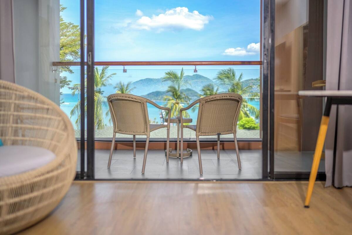 Een balkon met rieten stoelen, een kleine tafel en uitzicht op de oceaan en palmbomen.