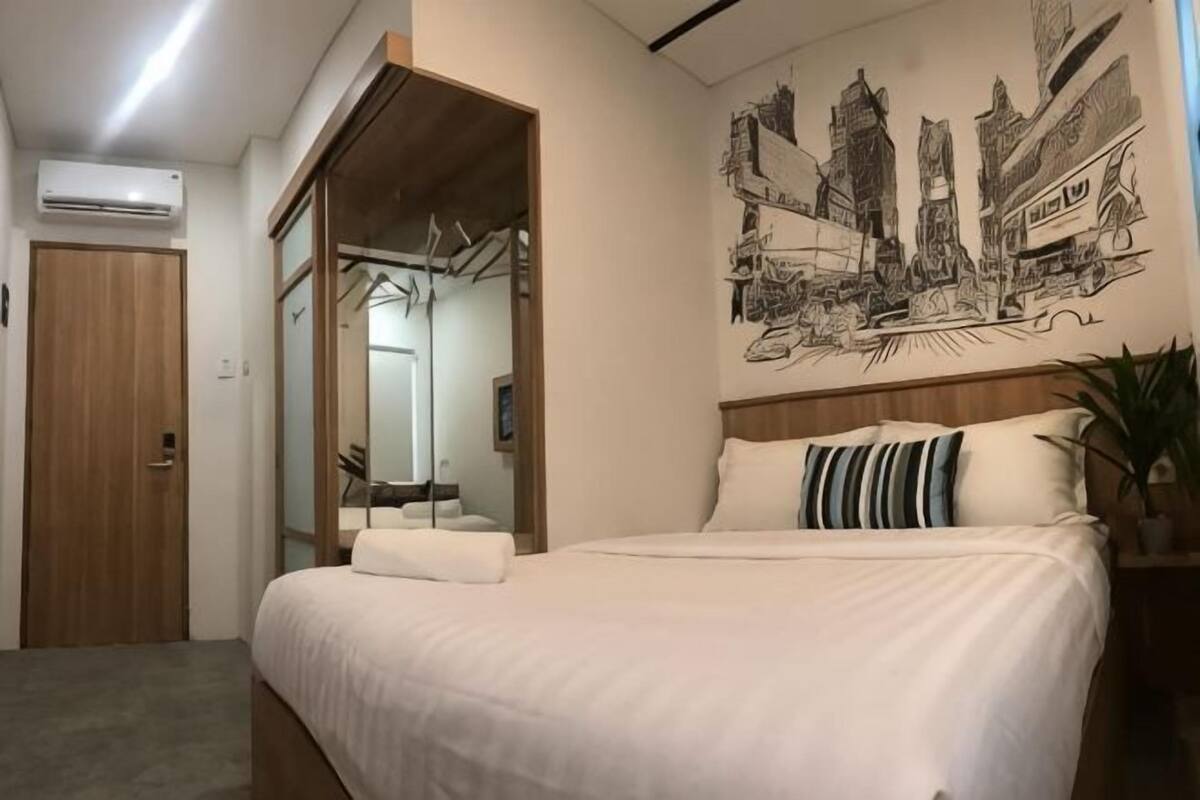 A bedroom with a bed, a wooden door, a mirror, and a cityscape wall art.