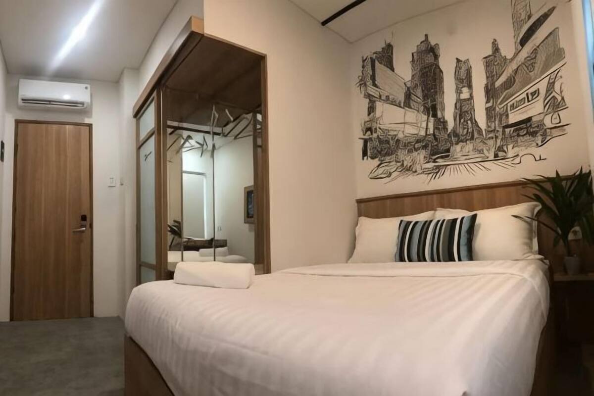 A bedroom with a bed, a wooden door, a mirror, and a cityscape artwork on the wall.