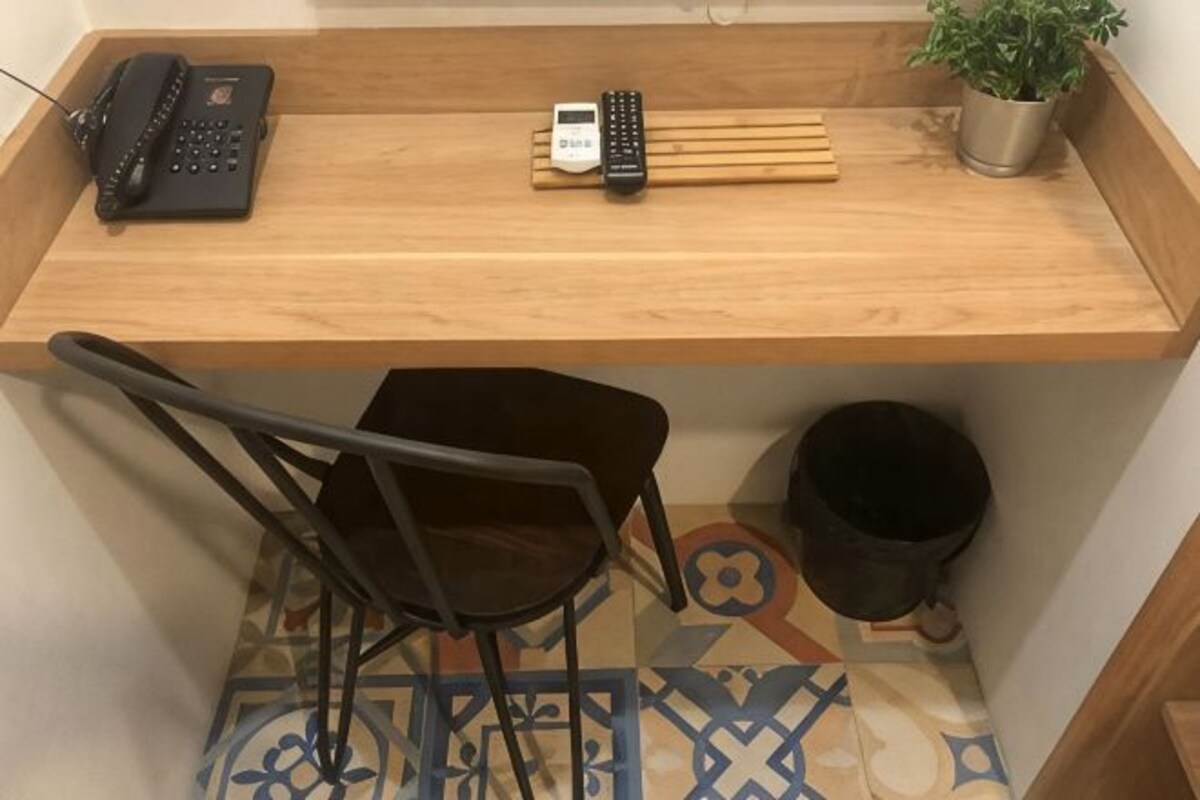 A wooden desk with a telephone, remote control, and a potted plant. Below the desk, there is a chair and a trash bin.