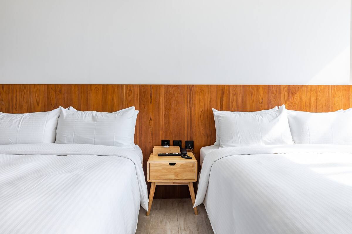 A hotel room with two beds, a wooden headboard, a nightstand with a drawer, and a wall-mounted power outlet.