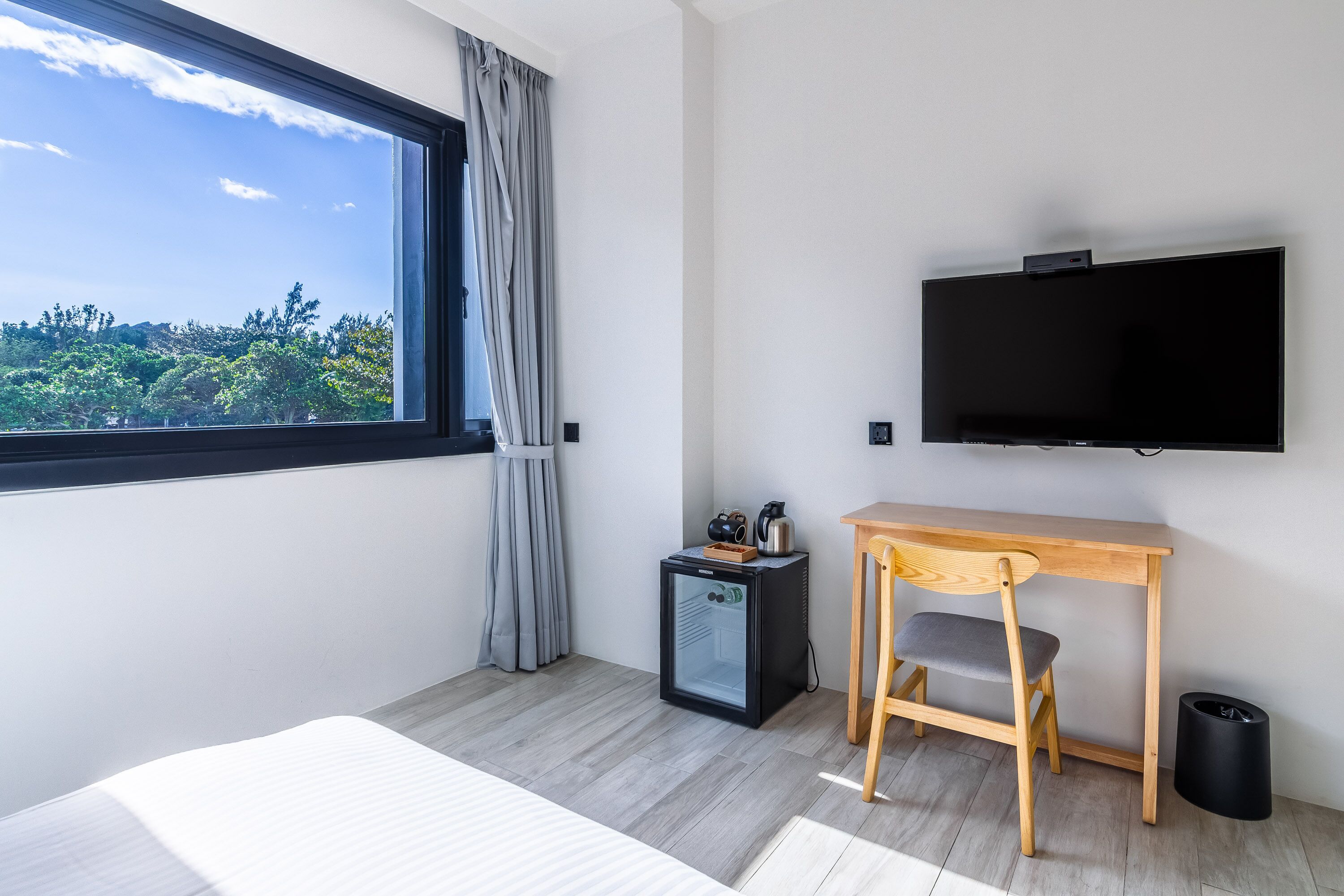 Una habitación de hotel moderna con un ventanal grande, un televisor de pantalla plana, un refrigerador pequeño, un escritorio de madera y una silla.