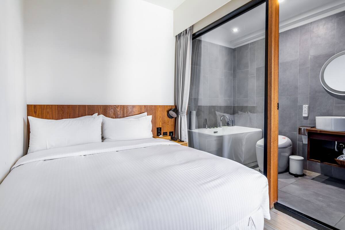 A modern hotel room with a large bed, a wooden headboard, a glass shower enclosure, a sink, and a mirror.