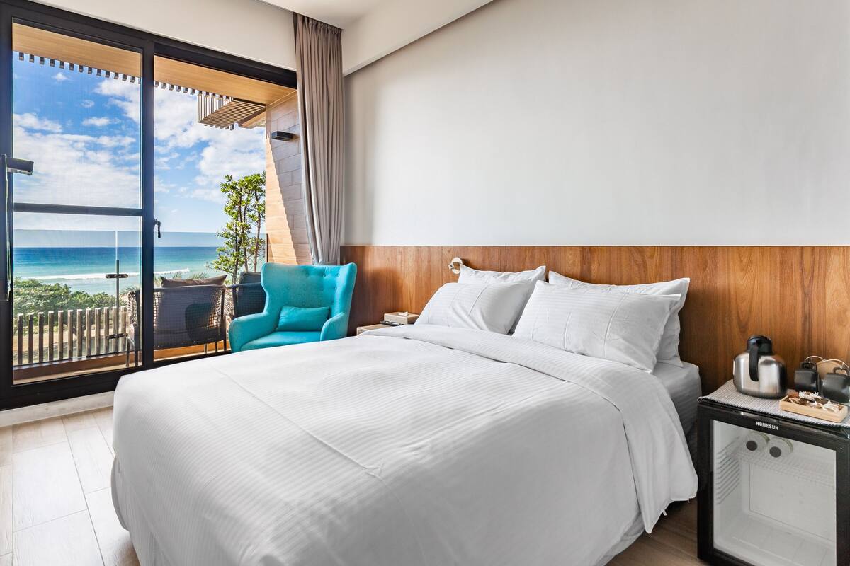 A hotel room with a large bed, a nightstand, a chair, and a view of the ocean.