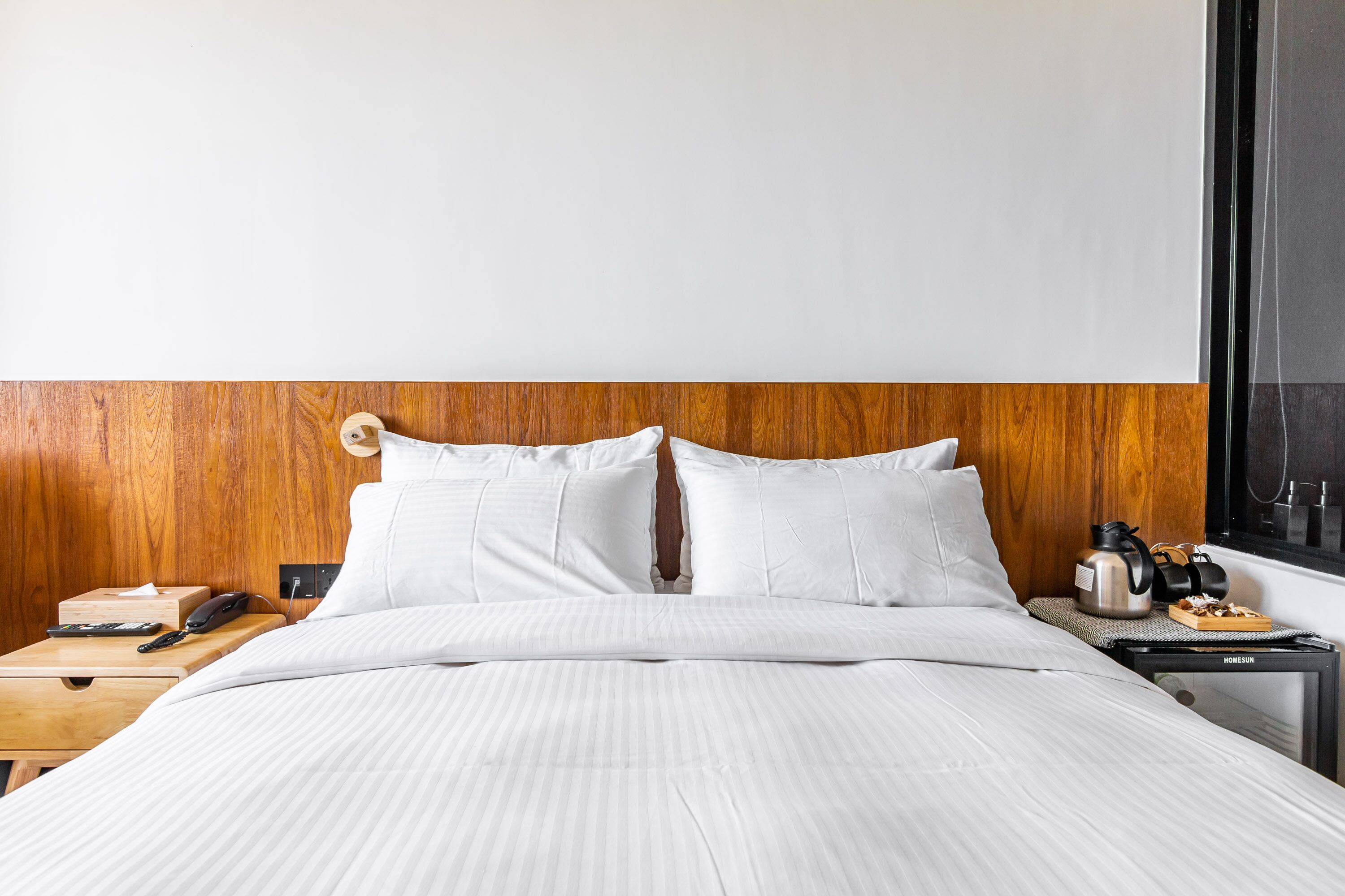 A neatly made bed with white linens, a wooden headboard, a bedside table with a phone, remote, and a kettle.
