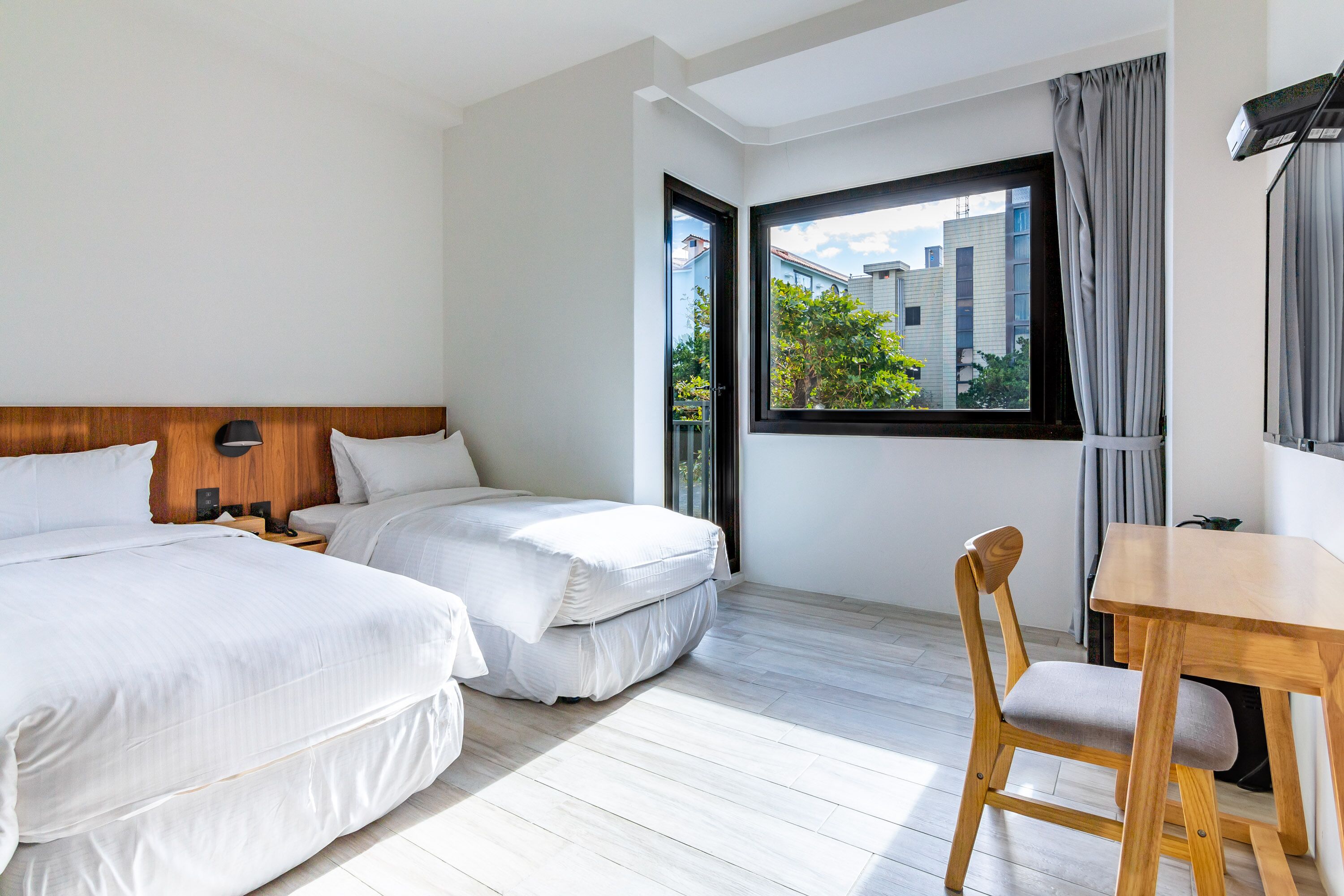 Een hotelkamer met twee bedden, een houten hoofdbord, een houten eettafel en uitzicht op gebouwen en bomen door een raam.