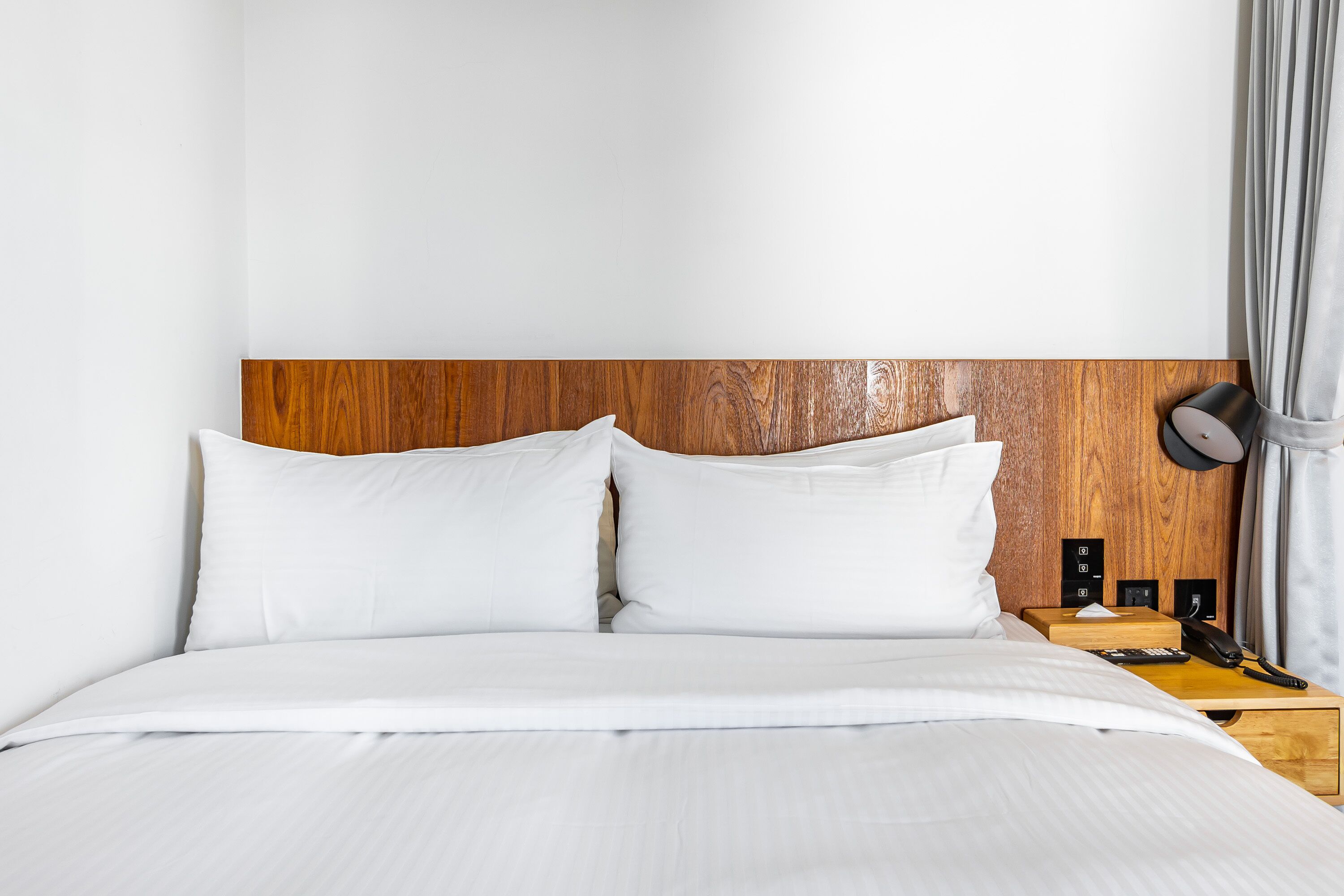 A neatly made bed with white linens, a wooden headboard, a bedside table with a lamp, and a telephone.