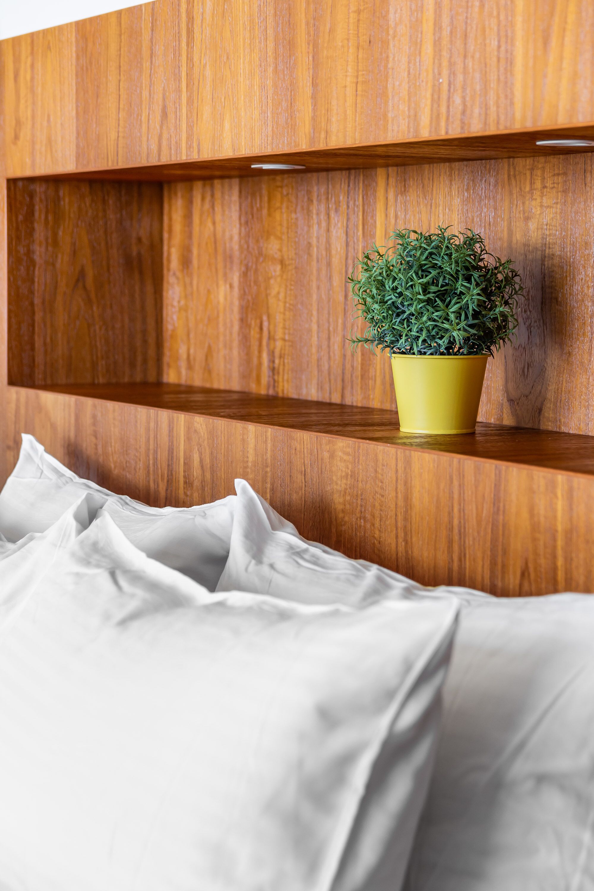 A wooden shelf with a potted plant and a neatly made bed with white bedding.