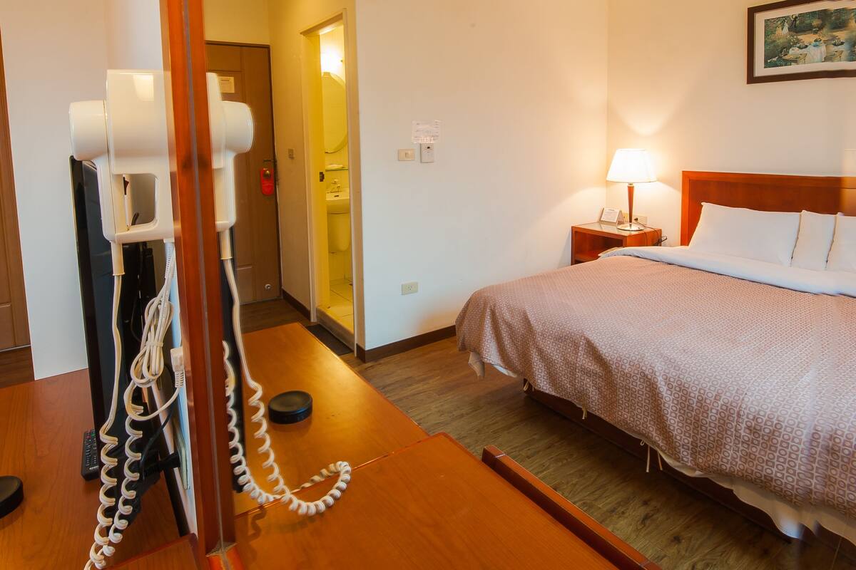 A hotel room with a wooden bed, a bedside table with a lamp, a telephone, and a door leading to a bathroom.