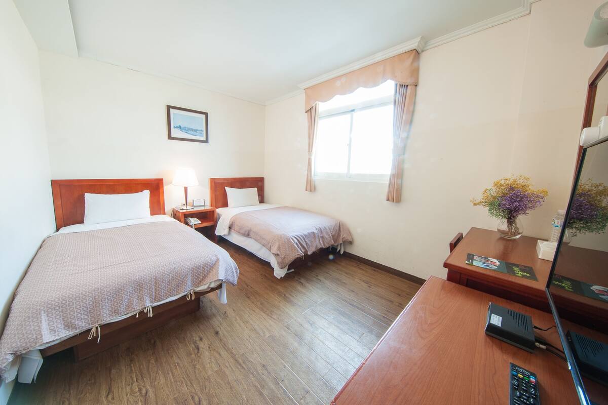 A hotel room with two beds, a desk with a computer, a telephone, and a window with curtains.