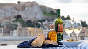 A table with bread, olives, a bottle of olive oil, and two glasses of white wine, set against a backdrop of a historic building and a cliff. - Hapimag Resort Athens (Athens)