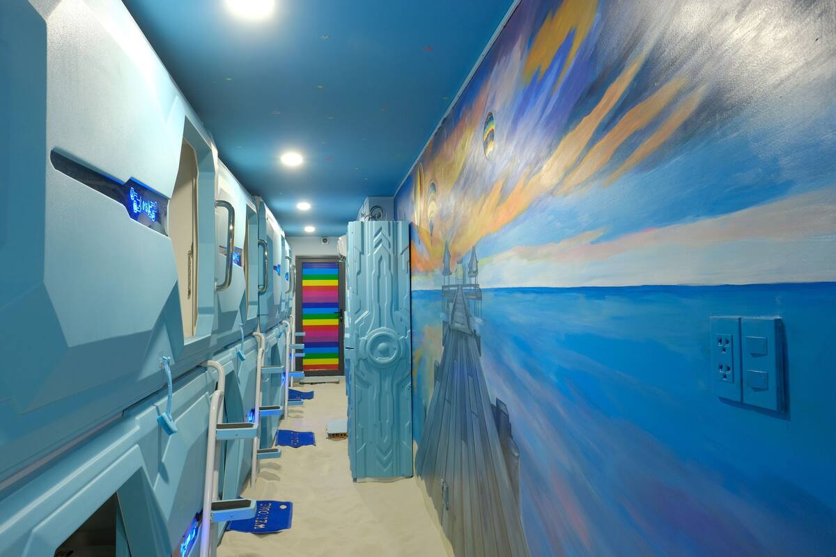 Un couloir avec un mur bleu et une fresque représentant un navire. Il y a des portes sur le côté gauche et une porte colorée au centre.
