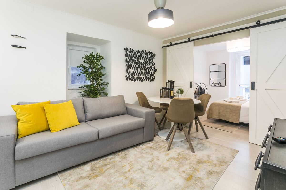 A modern living room with a grey sofa, yellow cushions, a dining table with chairs, a large plant, and a kitchen area with a sliding door.