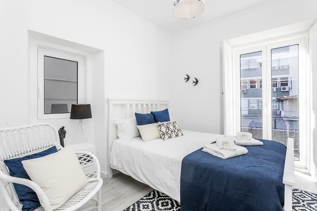 A bedroom with a white bed, a white chair, a patterned rug, and a view of buildings through a window.