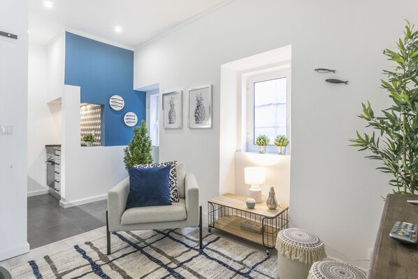 A modern living room with a blue accent wall, a sofa with a patterned pillow, a coffee table with decorative items, and a large window with a view of greenery.