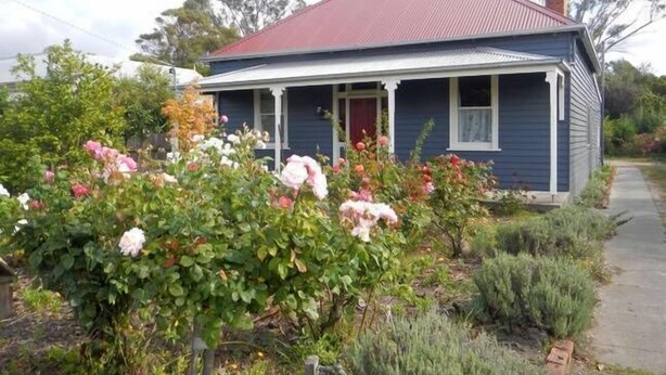 Une maison à deux étages, avec un toit rouge et une cheminée, entourée d’un jardin où poussent différentes plantes et fleurs.