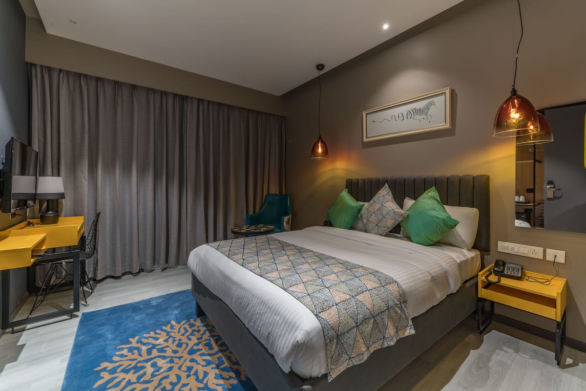 A modern hotel room with a large bed, a desk with a lamp, a chair, and a blue patterned rug.